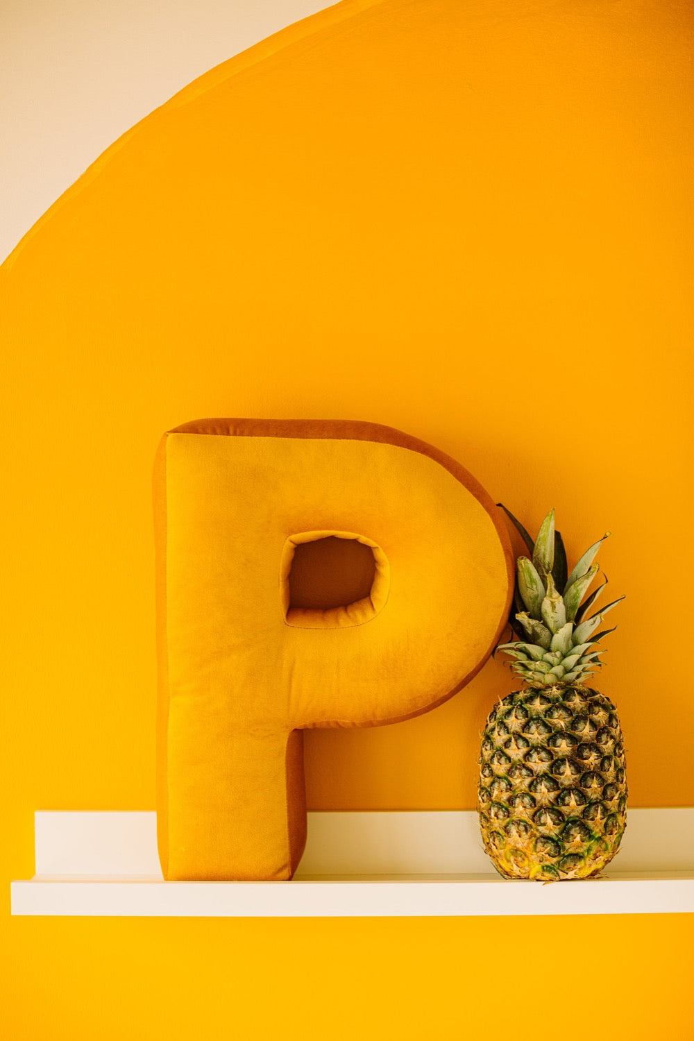 Na tle żółtej ściany na półce stoi welurowa poduszka w kształcie literki P. Obok literki jest ananas.