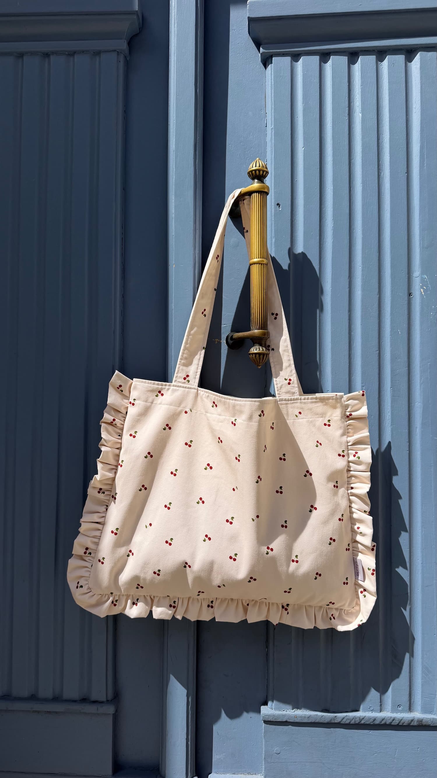 Bawełniana torba Szoperka z nadrukiem wiśni i falbanką od Bettys Home. Torba tote bag wisząca na niebieskich drzwiach