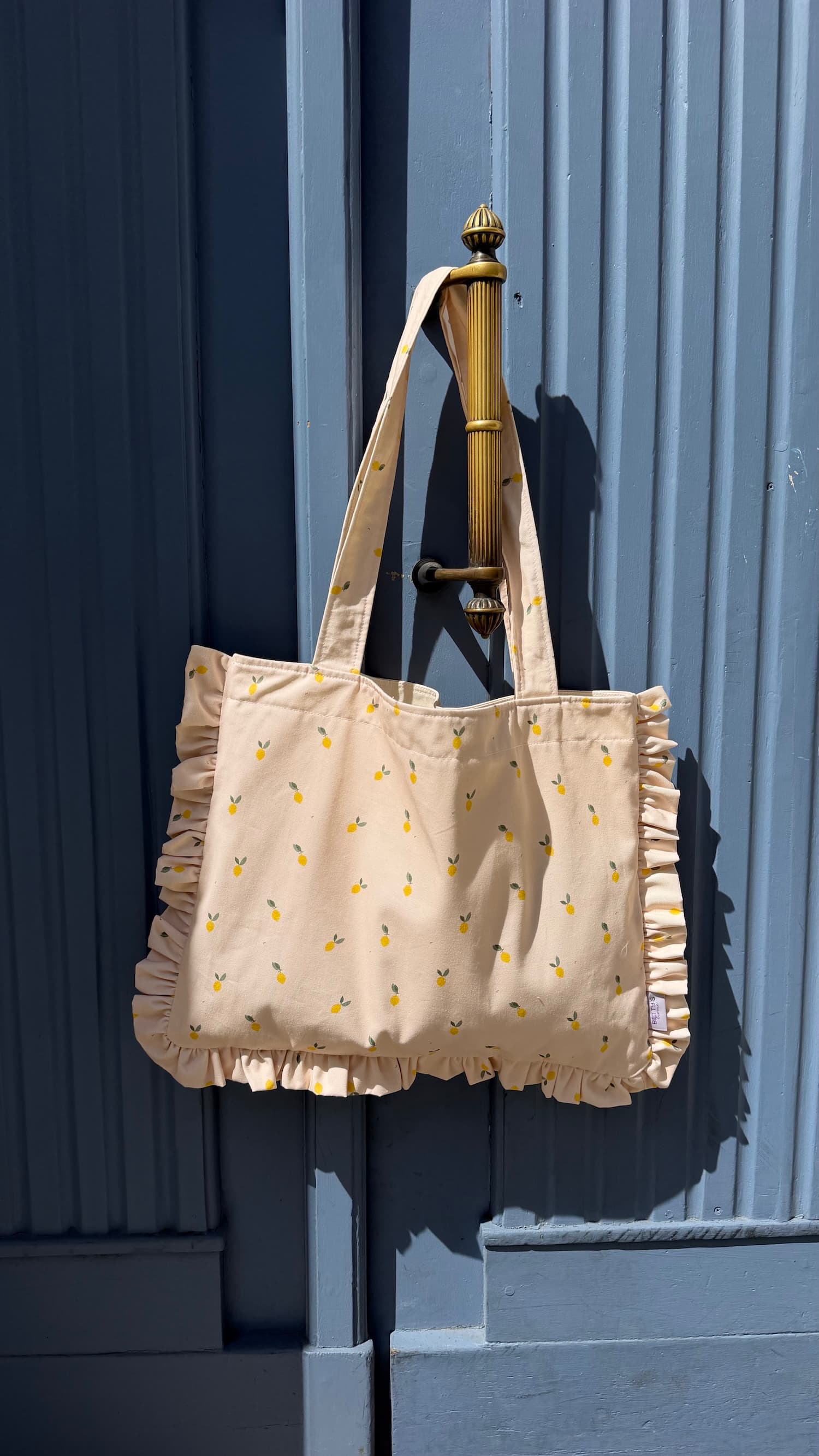 Bawełniana torba Szoperka z nadrukiem cytryn i falbanką od Bettys Home. Torba tote bag wisząca na niebieskich drzwiach