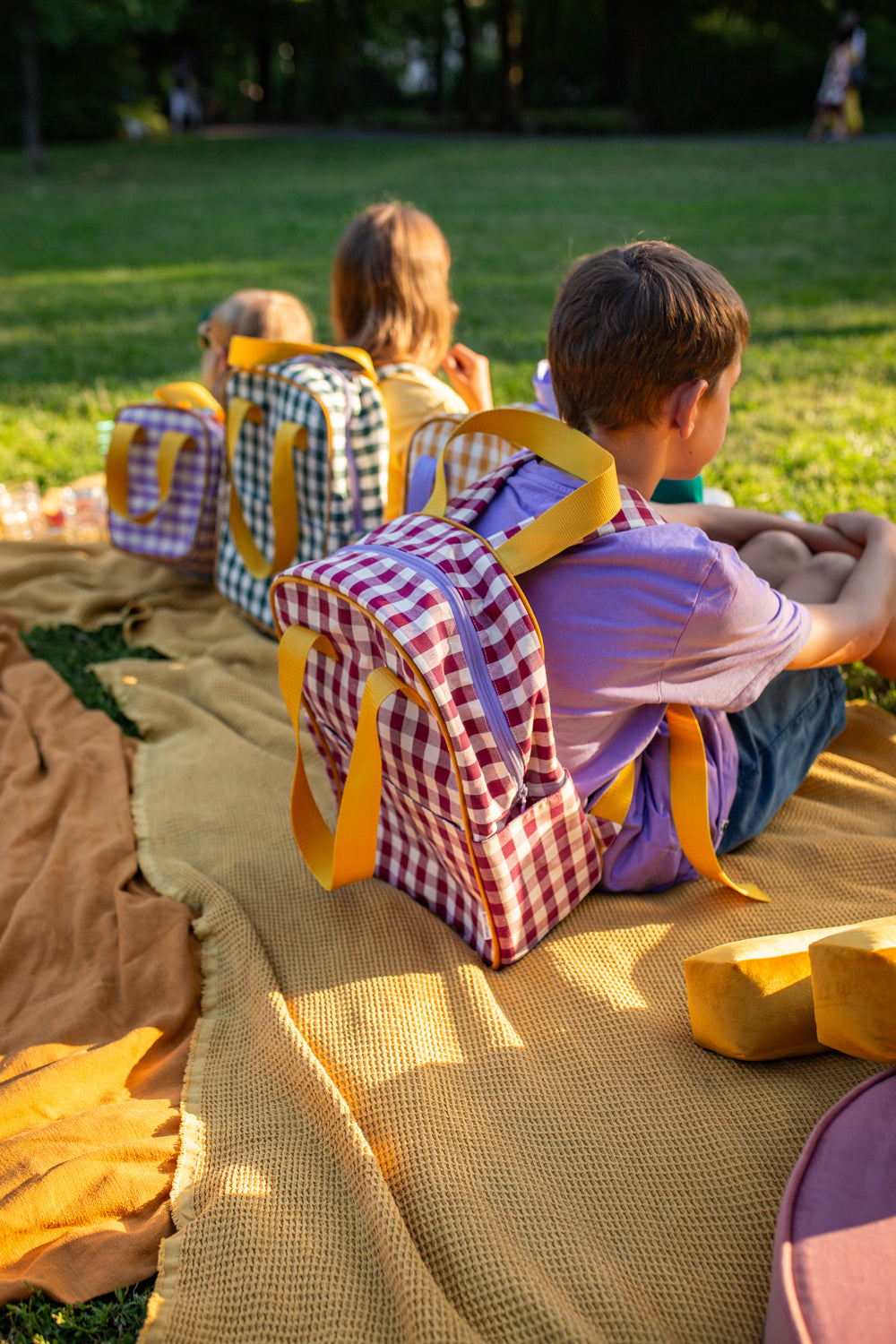 dzieci siedzące na kocu na trawie z plecakami w kratkę w kolorze czerwonym i zielonym od Bettys Home, plecak dla przedszkolaka
