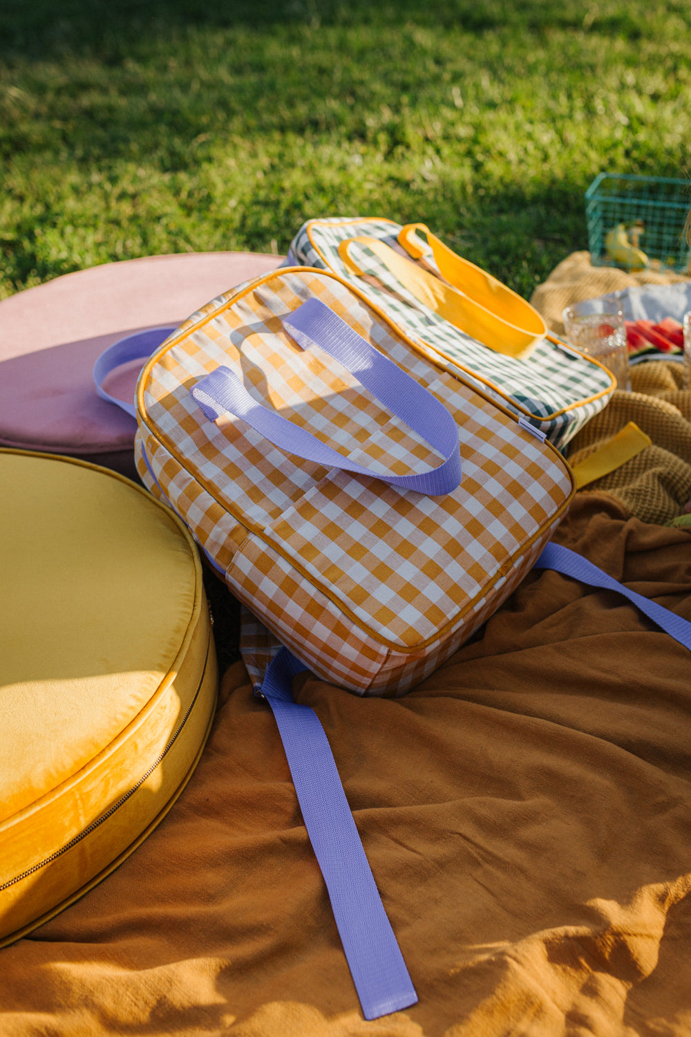 plecak w kratkę żółty i plecak zielony w kratkę od Bettys Home leżące na kocu w trakcie pikniku obok welurowej pufy żółtej 