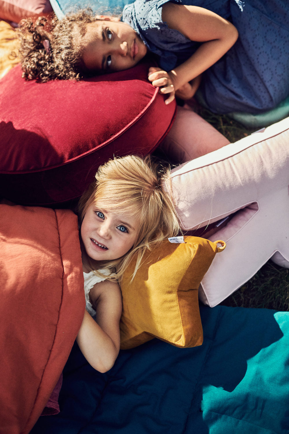 dwie dziewczynki leżące pomiędzy poduszkami. Jedna z nich ma głowę opartą na małe poduszce gwiazdce w kolorze żółtym 