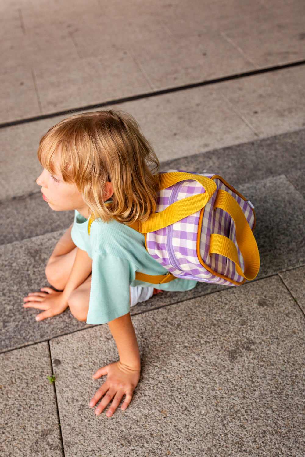 chłopiec siedzący na chodniku z małym fioletowym plecakiem w kratkę od Bettys Home, plecak szkolny, plecak dla przedszkolaka