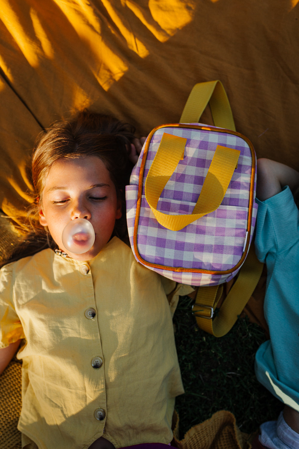 Dziewczynka robiąca balona z gumy obok niej mały plecak fioletowy w kratkę od Bettys Home, plecak do szkoły dla dziewczynki 