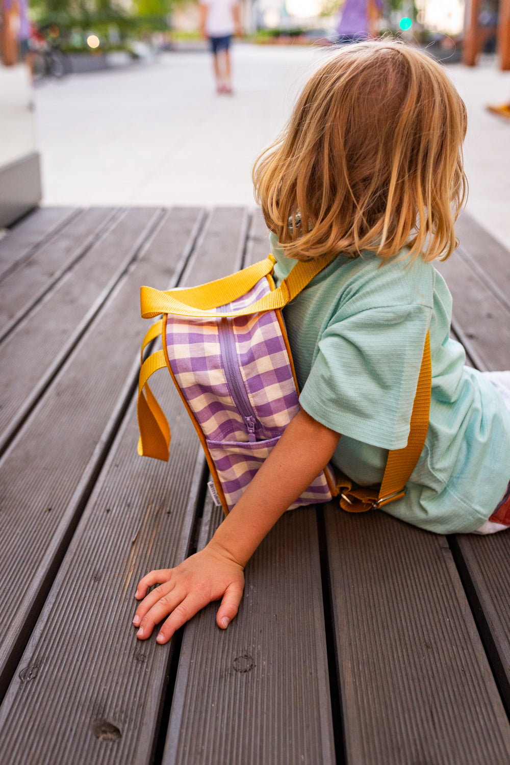 chłopiec siedzący na drewnianym tarasie, ma mały plecak w kratkę fioletowy od Bettys Home, plecak do szkoły dla chłopca 