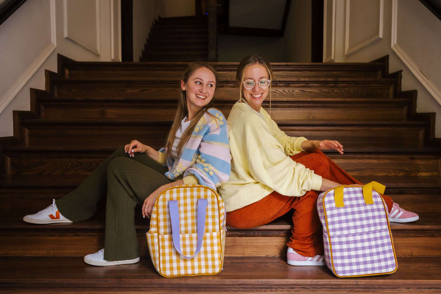 dwie uśmiechnięte dziewczyny siedzące na schodach a przed nimi stoją dwa plecaki w kratkę w kolorze fioletowym i żółtym 
