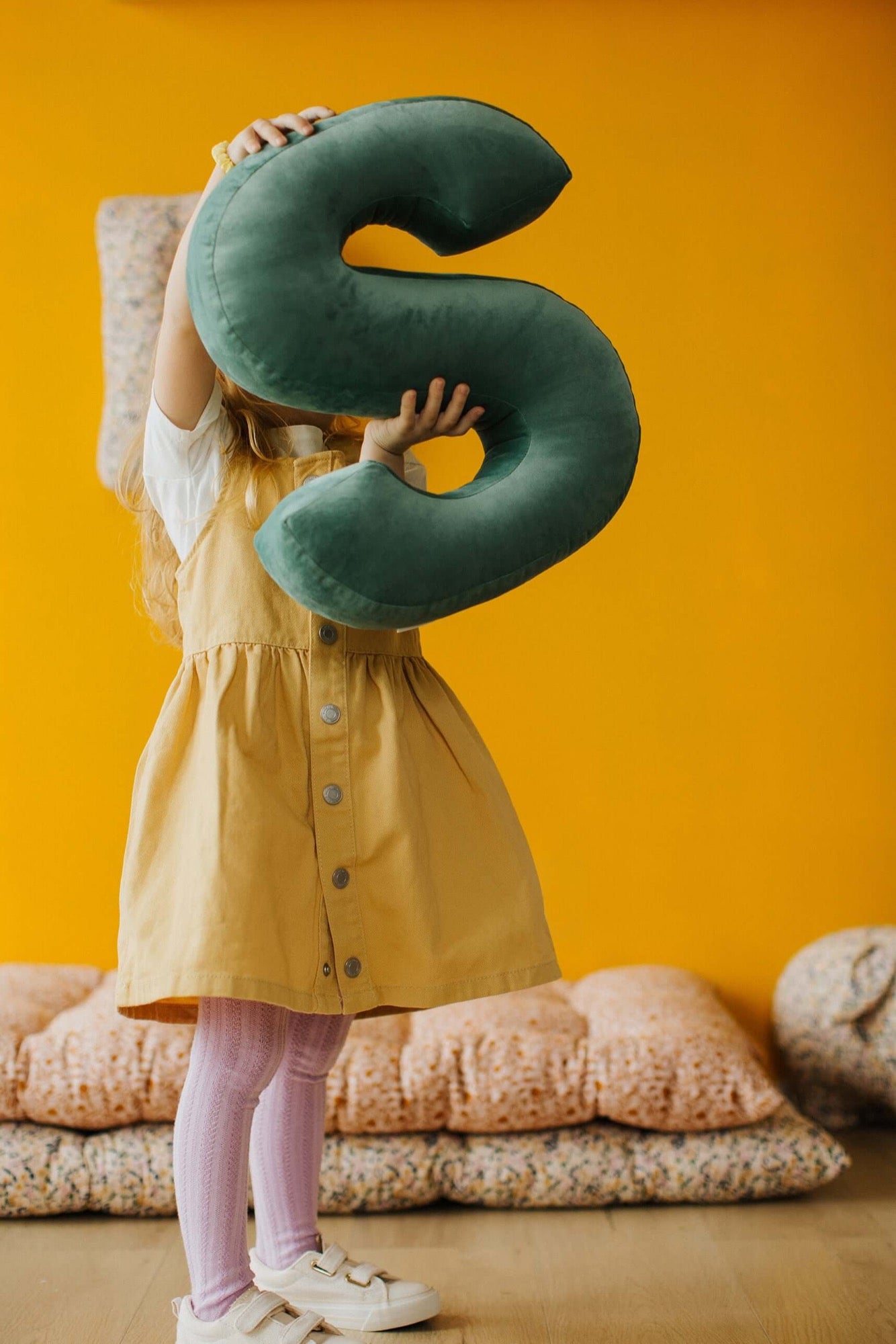 Dziewczynka prezentuje poduszkę literkę welurową S zieloną od Bettys Home na tle żółtej ściany 