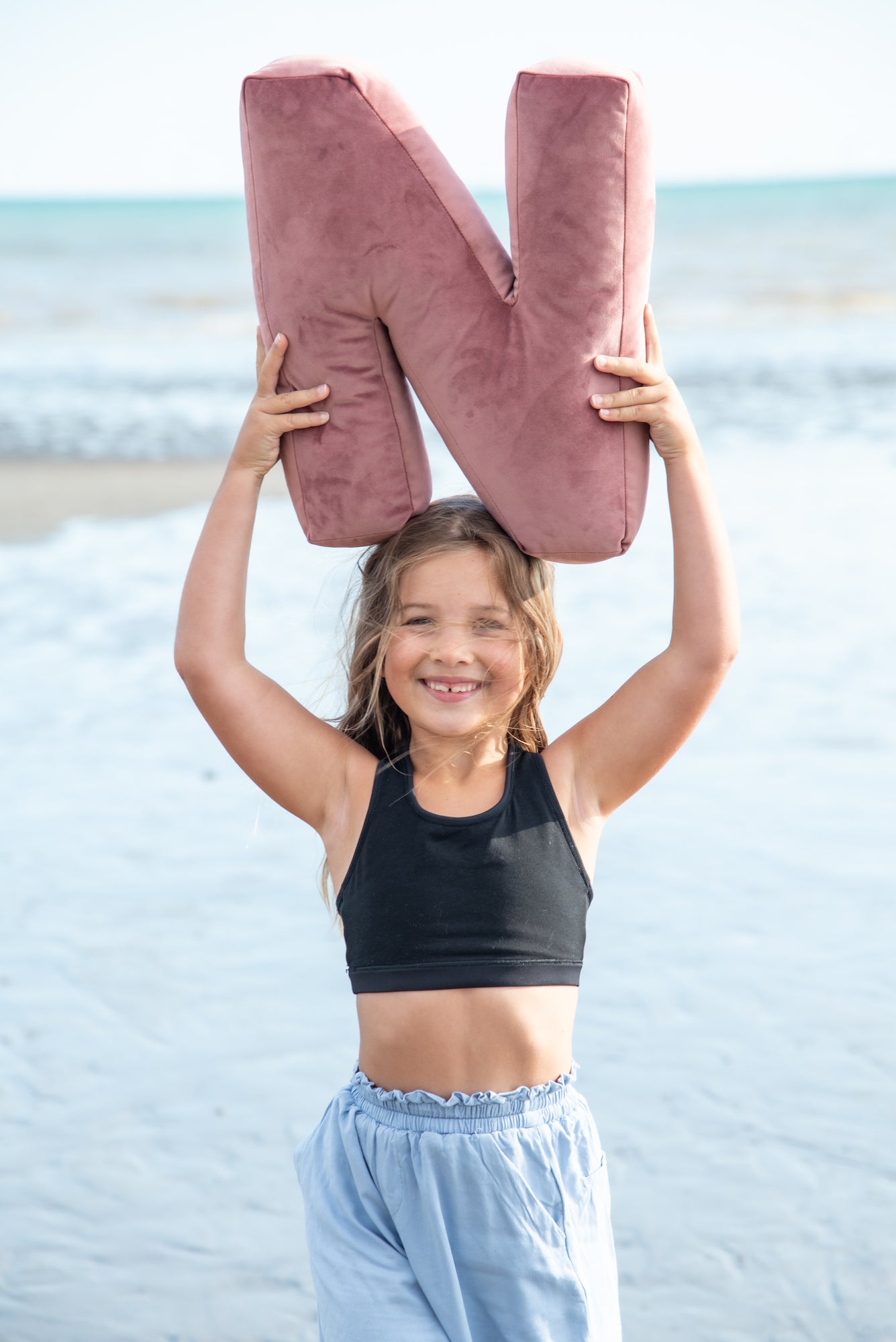 urocza dziewczynka na plaży trzyma nad głową różową poduszkę literkę N od Bettys Home