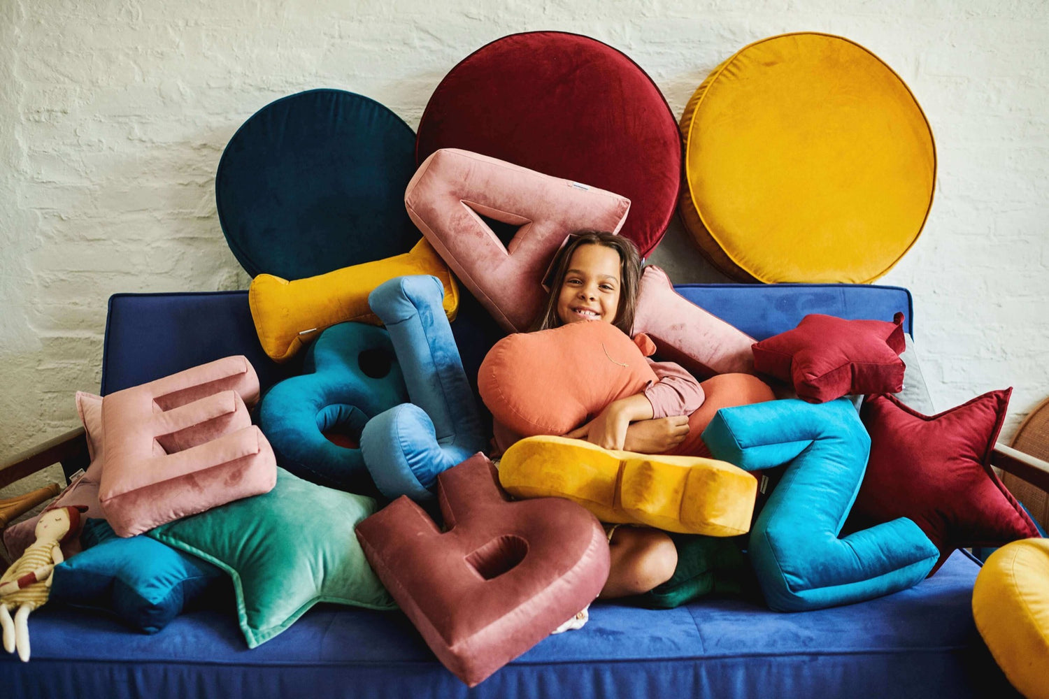 dziewczynka siedząca na kanapie, przykryta poduszkami literkami welurowymi i pufami welurowymi
