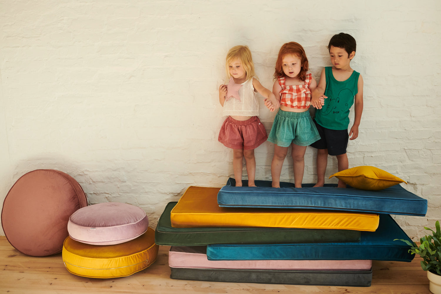 Kids standing on top of stacked mattresses in different colours. Next to them are round velvet poufs
