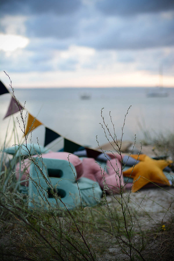 Na tle morza poduszki w kształcie literek leżą na plaży, a nad nimi wisi girlanda. 
