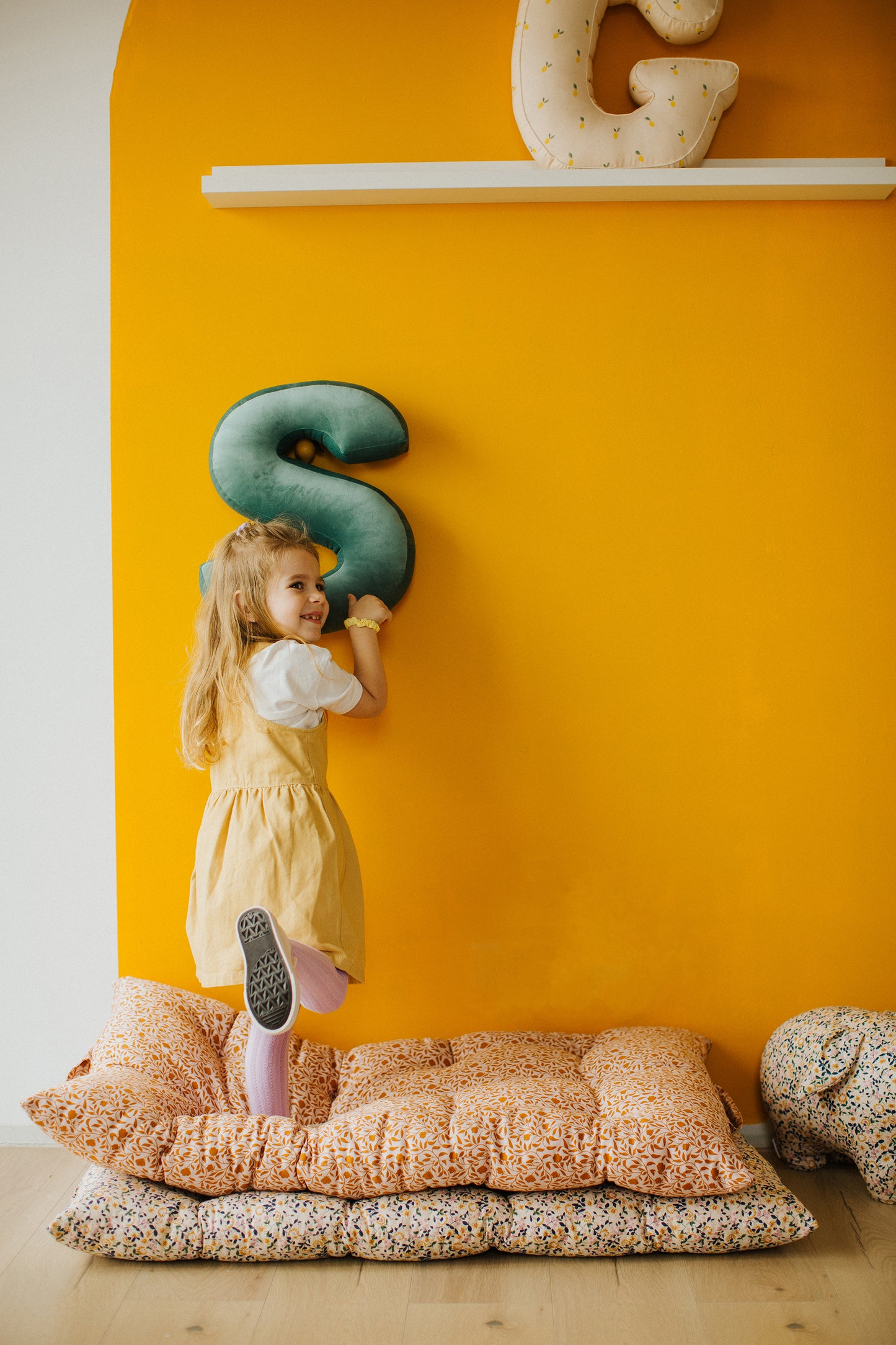 Mała dziewczynka trzyma w rękach poduszkę w kształcie S. Jest w trakcie zabawy poduszkami literkami. 