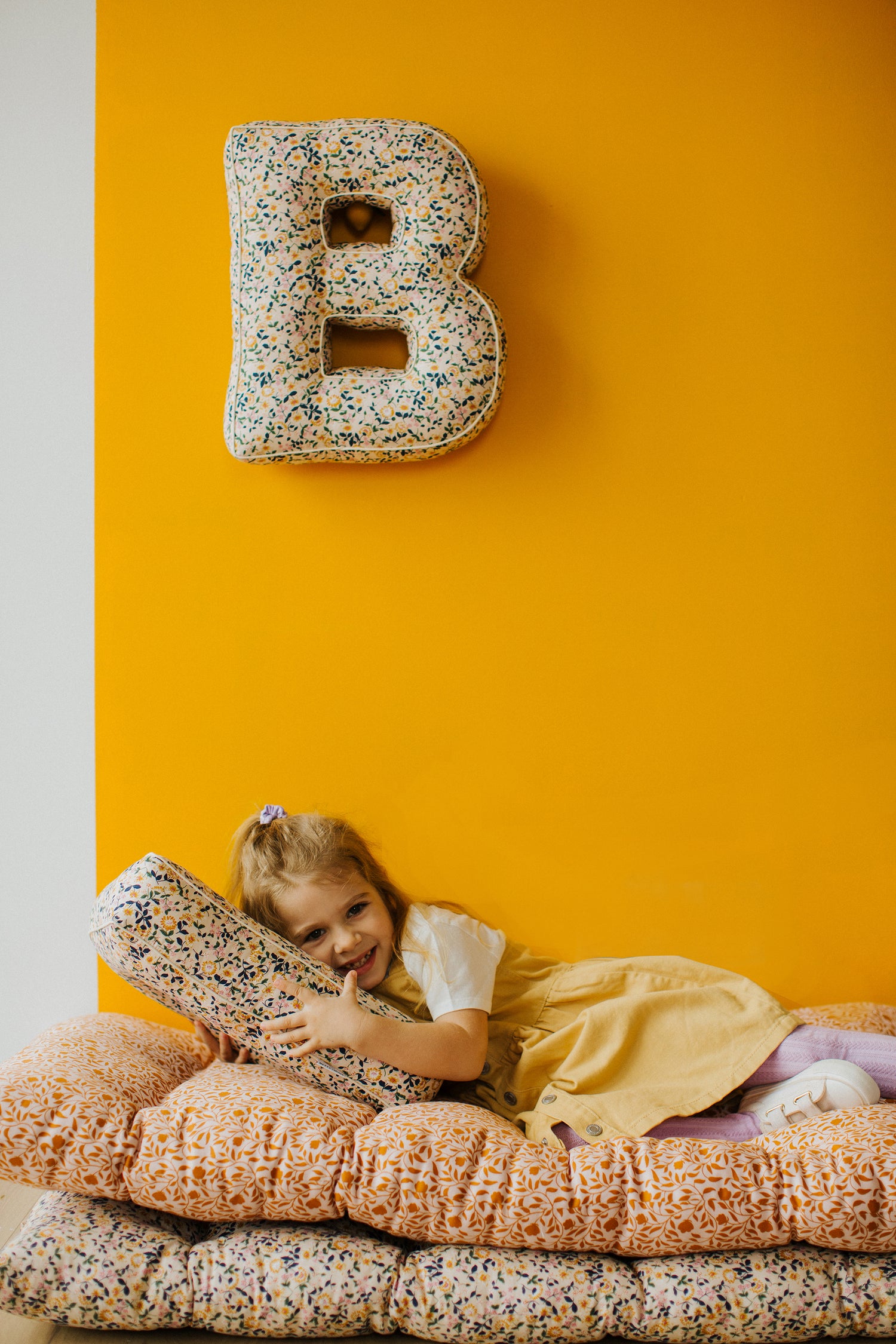 Dziewczynka leżąca na miękkim materacu Betty’s Home. Nad nią na ścianie wisi poduszka literka w kwiatki w kształcie B. 