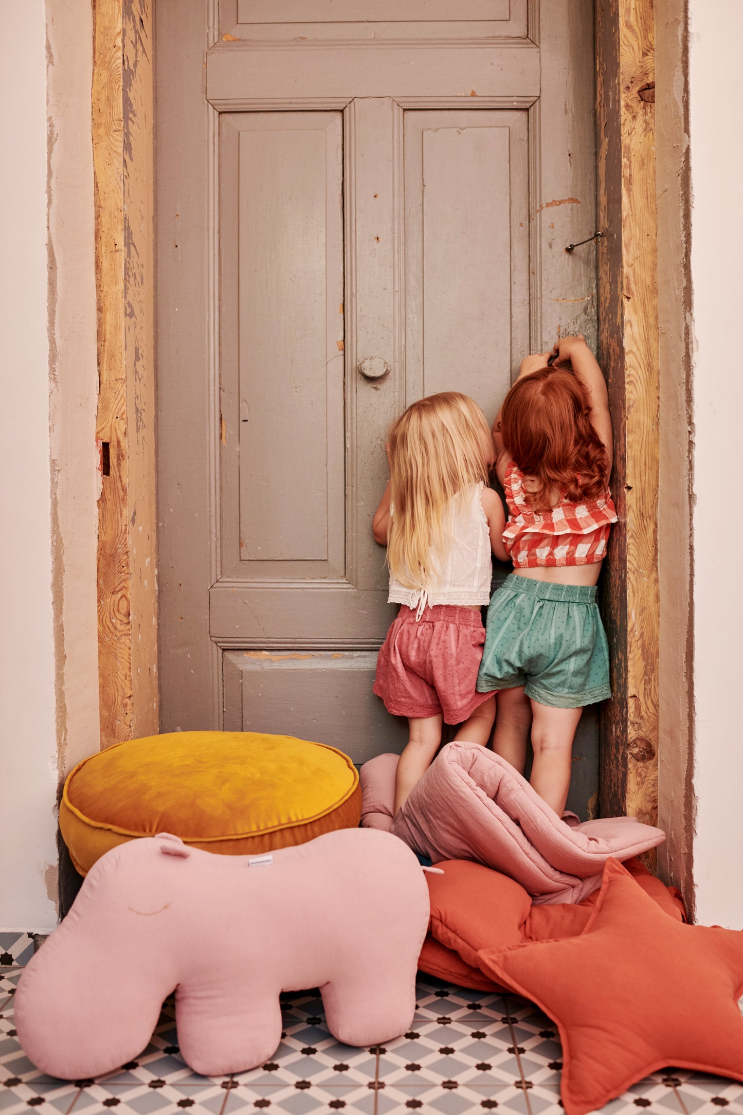 Two girls standing on rolled pink and red floormats are trying to open beige door. Next to them is yellow pouf and pink hippo cushion. 